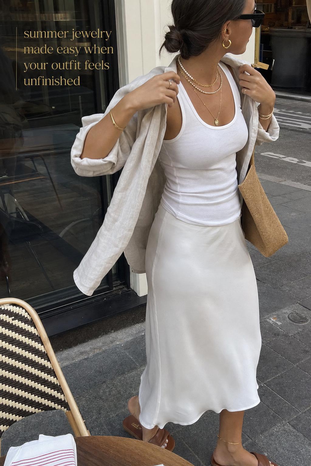 Candid street-style photo of layered summer jewelry on a woman adjusting a linen shirt in warm daylight
