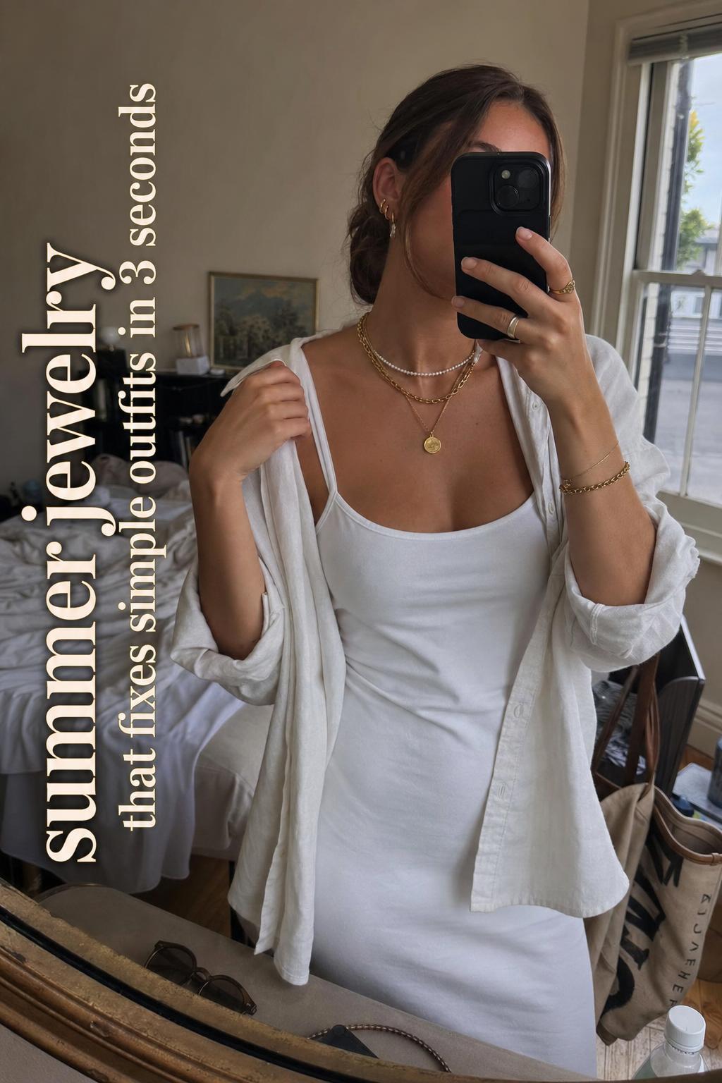 Mirror selfie styling summer jewelry with layered chains and bracelets in a casual messy bedroom by window light