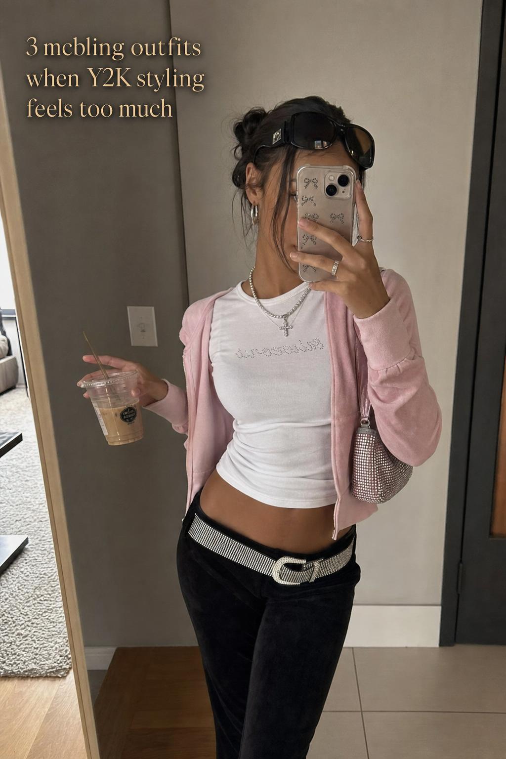 Young woman in blush velour hoodie and rhinestone belt showing modern mcbling outfits in a candid hallway mirror selfie.