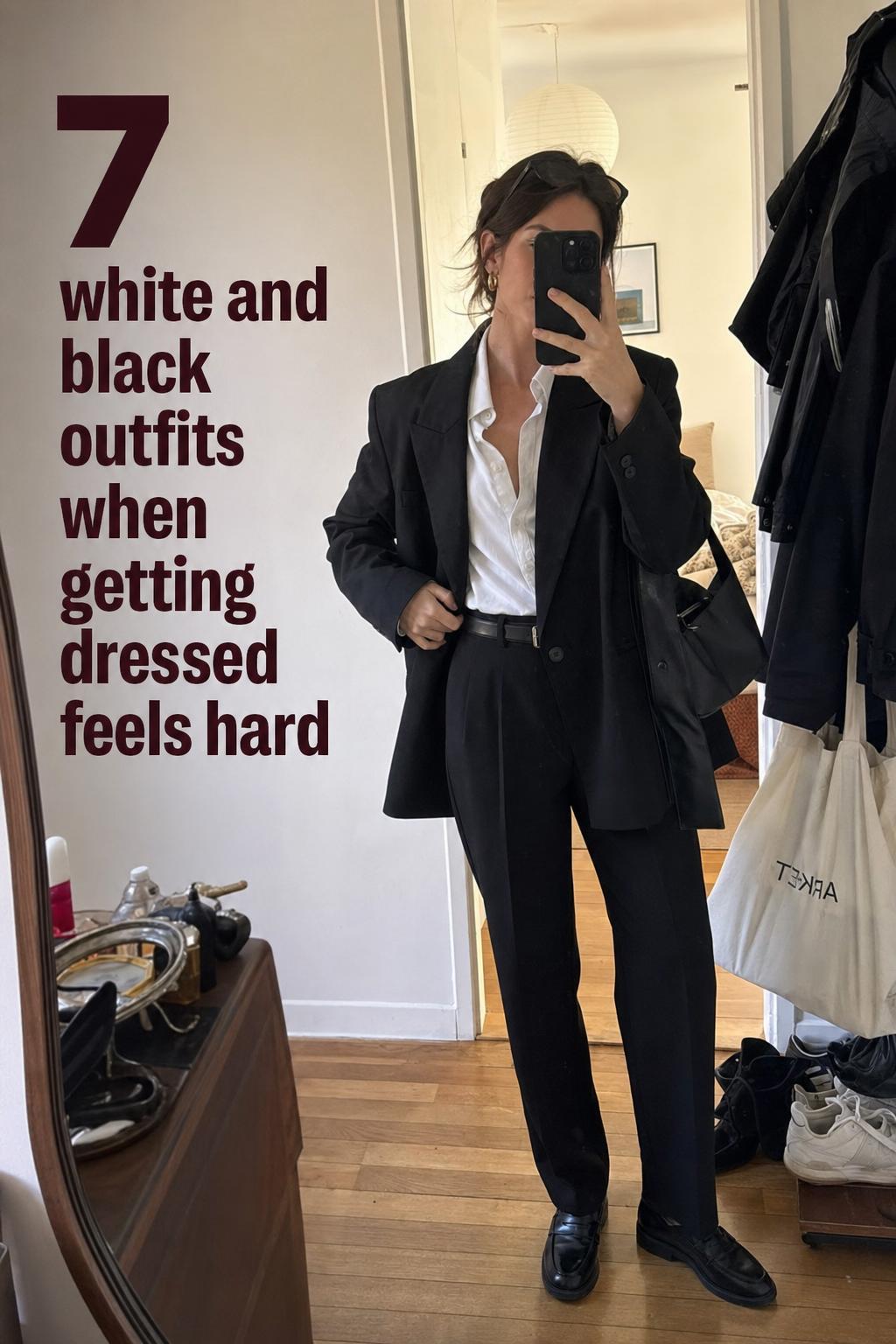 White and black outfits mirror selfie of a woman adjusting a black blazer over a white shirt in a lived-in apartment hallway